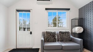 A modern living room with a grey leather sofa, two fluffy throw pillows, a black and white patterned wall, a black metal shelf with a rolled-up towel, and a black doormat. - Luxe Lodges on Lake Erie ~ Bunkie ~ Beach Access (Port Dover)