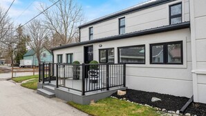 A modern two-story house with a balcony, a small porch, and a well-maintained lawn. - Luxe Lodges on Lake Erie ~ Emerald ~ Beach + Patio (Port Dover)