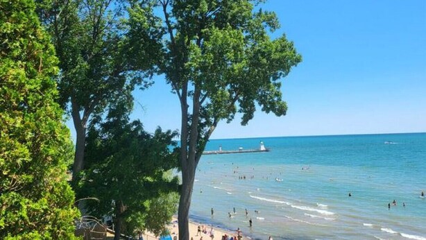 A beach with clear skies, a lighthouse in the distance, and people swimming and relaxing on the shore. - Luxe Lodges on Lake Erie ~ Emerald ~ Beach + Patio (Port Dover)