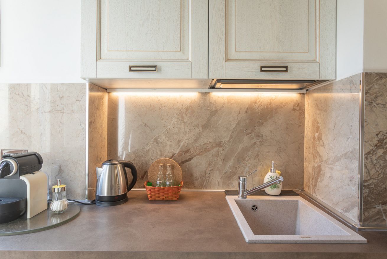 Una cocina con encimeras de mármol, cafetera, hervidor, una cesta con botellas y un lavabo.