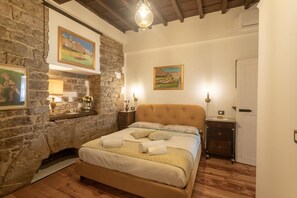 A bedroom with a stone wall, a bed with a headboard, a nightstand, a lamp, and a painting on the wall. - Old Bridge Boutique (Firenze)