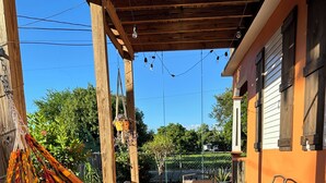 Eine Hängematte mit gestreifter Decke, eine Holz-Pergola, Gartenmöbel und Blick auf ein Haus mit Zaun und Grünpflanzen.