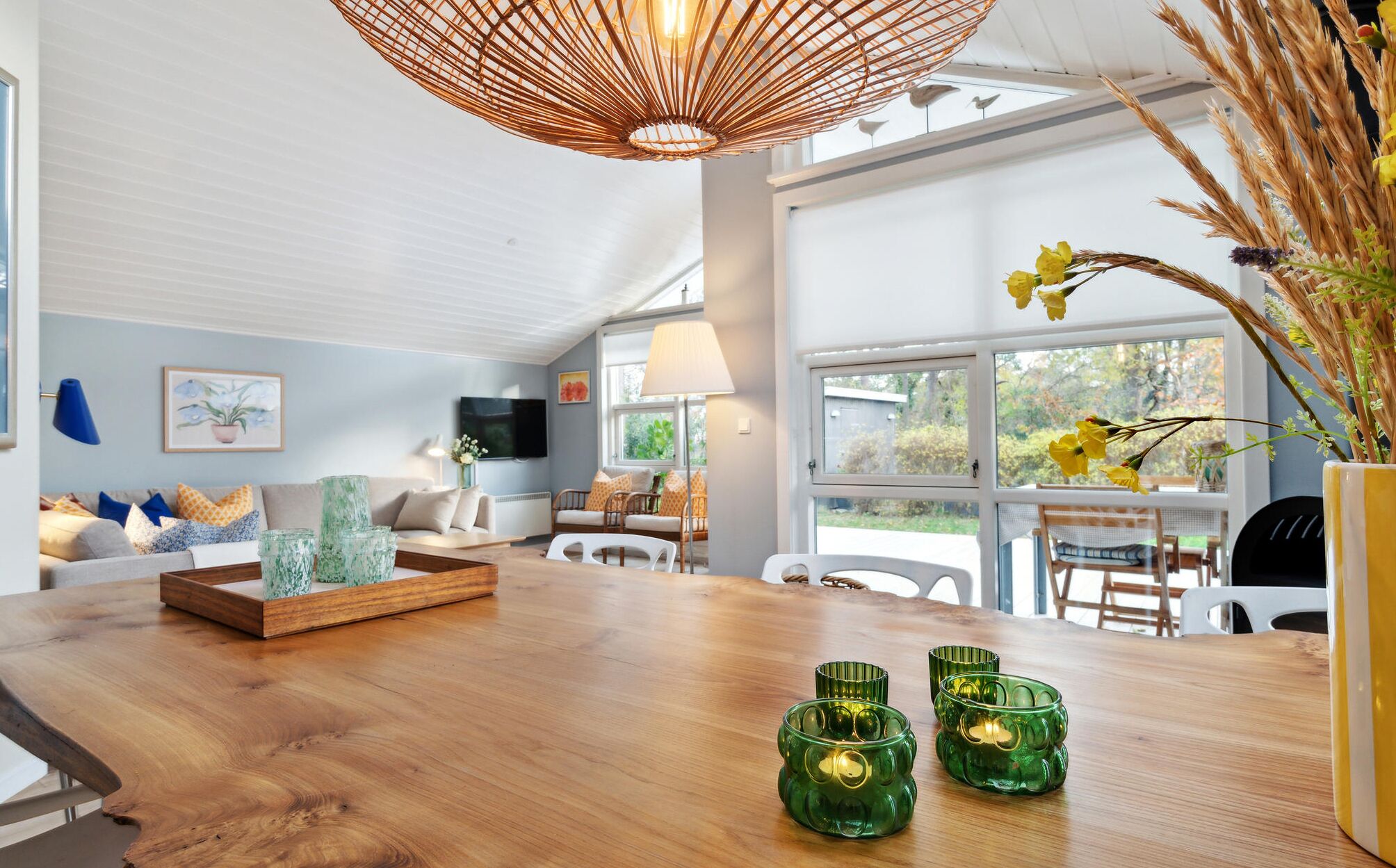 A modern living room with a wooden dining table, green glass candle holders, and a large window with a view of greenery.