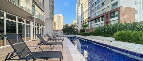 A modern outdoor swimming pool with lounge chairs, surrounded by high-rise buildings and greenery.