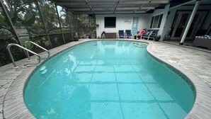 Una piscina con borde de baldosas, un área techada con sillas y una puerta al fondo.