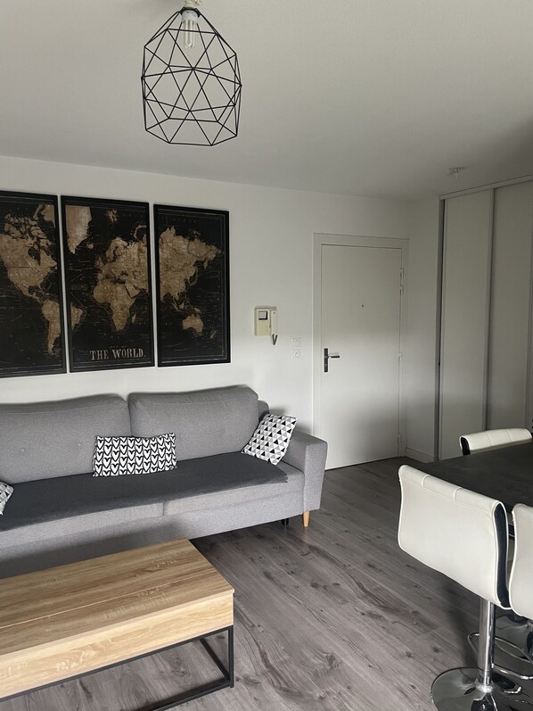 A modern living room with a grey sofa, a wooden coffee table, a dining table with white chairs, and a world map on the wall.