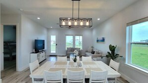 A spacious dining area with a large table and chairs, a television in the background, and a view of the outdoors through large windows.