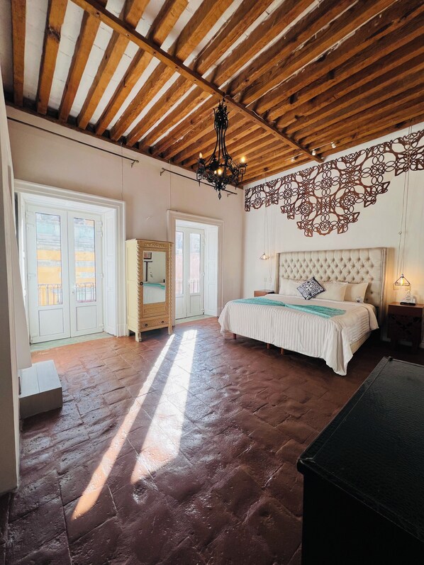 Un dormitorio con techo de madera, una cama con cabecera capitonada, una araña y un elemento decorativo en la pared.