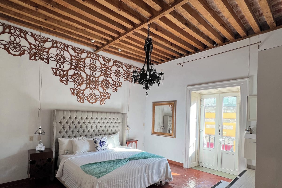 Ein Schlafzimmer mit Holzbalkendecke, einem Bett mit einer gepolsterten Kopfteil und einem Kronleuchter, sowie einem Blick ins Freie durch eine Tür.
