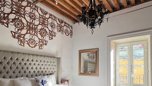 Une chambre à coucher avec une tête de lit capitonnée, un grand lit recouvert d’une couvre-lit à rayures, un lustre suspendu, un élément décoratif mural et une porte donnant sur l’extérieur.