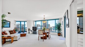 A spacious living room with a white sofa, a dining table, and chairs. There is a TV, a plant, and a large window offering a city view. - Stunning High-Floor Unit with Spectacular Ocean, Diamond Head, and Waikiki Views (Honolulu)