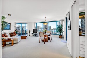 A spacious living room with a white sofa, a dining table, and chairs. There is a TV, a plant, and a large window offering a city view.