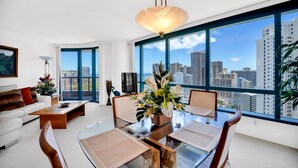 A modern living room with a glass dining table, white sofa, and large windows offering a city view. - Stunning High-Floor Unit with Spectacular Ocean, Diamond Head, and Waikiki Views (Honolulu)