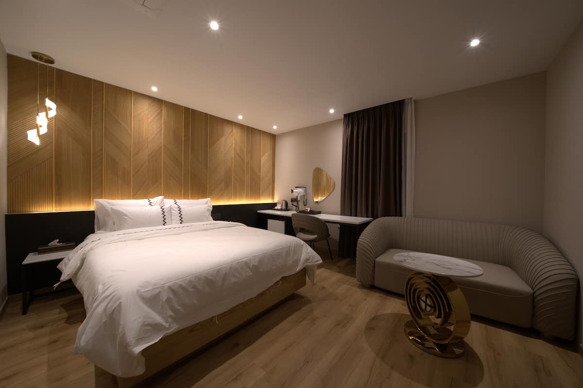 A modern hotel room with a large bed, a desk, a sofa, and a unique wall-mounted light fixture.