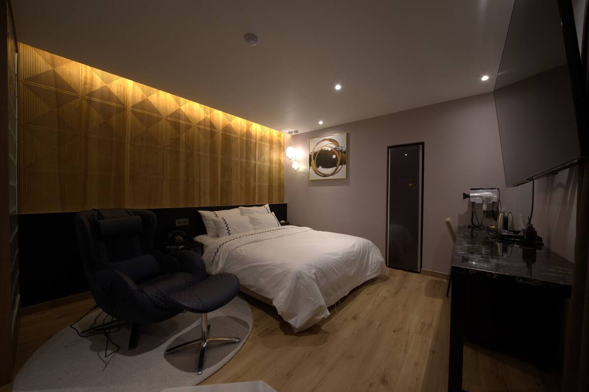 A modern bedroom with a bed, a massage chair, a desk, and a wall-mounted light fixture.