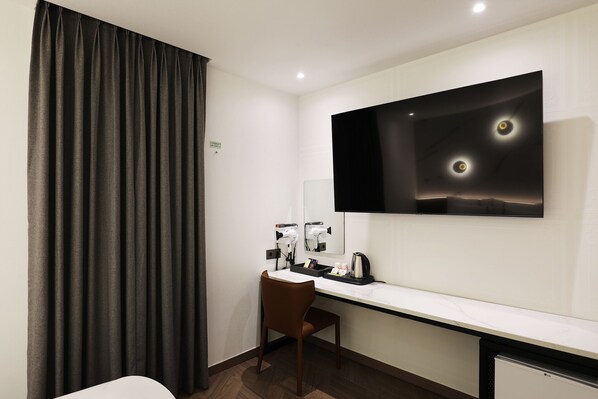 Habitación de hotel con escritorio, silla y un espejo grande. Hay un televisor de pantalla plana en la pared, y un secador de pelo enchufado.