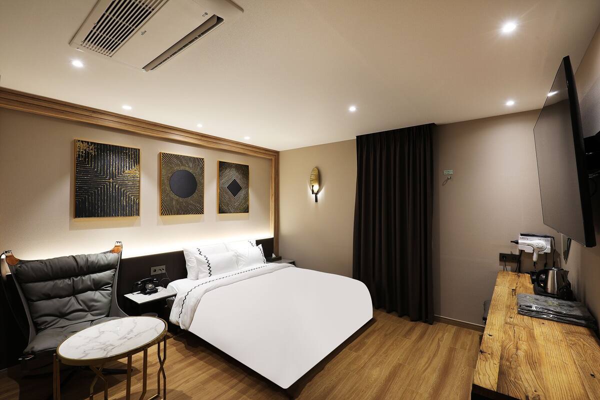 A hotel room with a bed, a seating area with a chair and a small table, a desk with a computer, a flat-screen TV, and artwork on the wall.