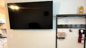 A living room with a flat-screen TV mounted on the wall, a dark blue ottoman, a wooden coffee table, and a metal shelf with hooks and a Texas flag. - Cuddly Critters Cottage - Alpacas, Goats, Emu's and YOU! (Blue Ridge)
