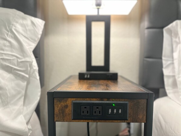 Ett litet bord som står vid sängen, med en grenuttag, en lampa och en spegel.