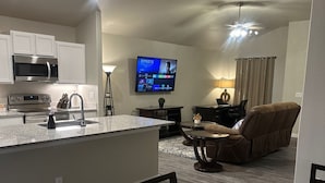 Una cocina moderna con microondas, estufa y lavaplatos incorporados, un comedor con mesa y sillas, y una sala de estar con sofá y televisor de pantalla plana reproduciendo un video musical.