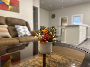 Un salon moderne avec une table basse en verre, un canapé agrémenté de coussins à motifs et, au fond, un espace cuisine.