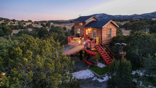 Une cabane en bois avec un toboggan rouge et des escaliers, entourée d’arbres et de montagnes.
