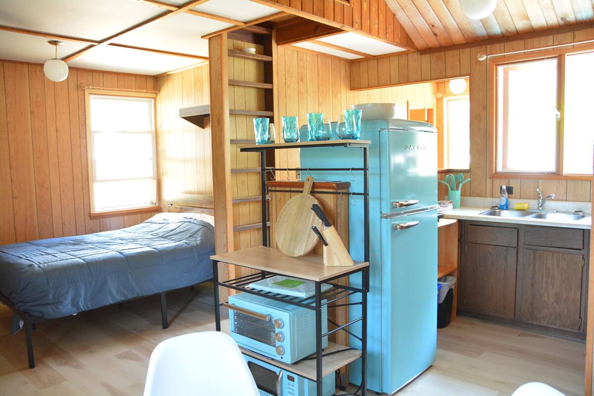 A compact kitchen with a blue refrigerator, a wooden shelf with kitchen utensils, and a bed in the adjacent room.