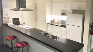 Una cocina moderna con gabinetes blancos, encimera negra, pileta de acero inoxidable, estufa a gas, microondas y refrigerador. Hay dos taburetes rojos con base de metal.