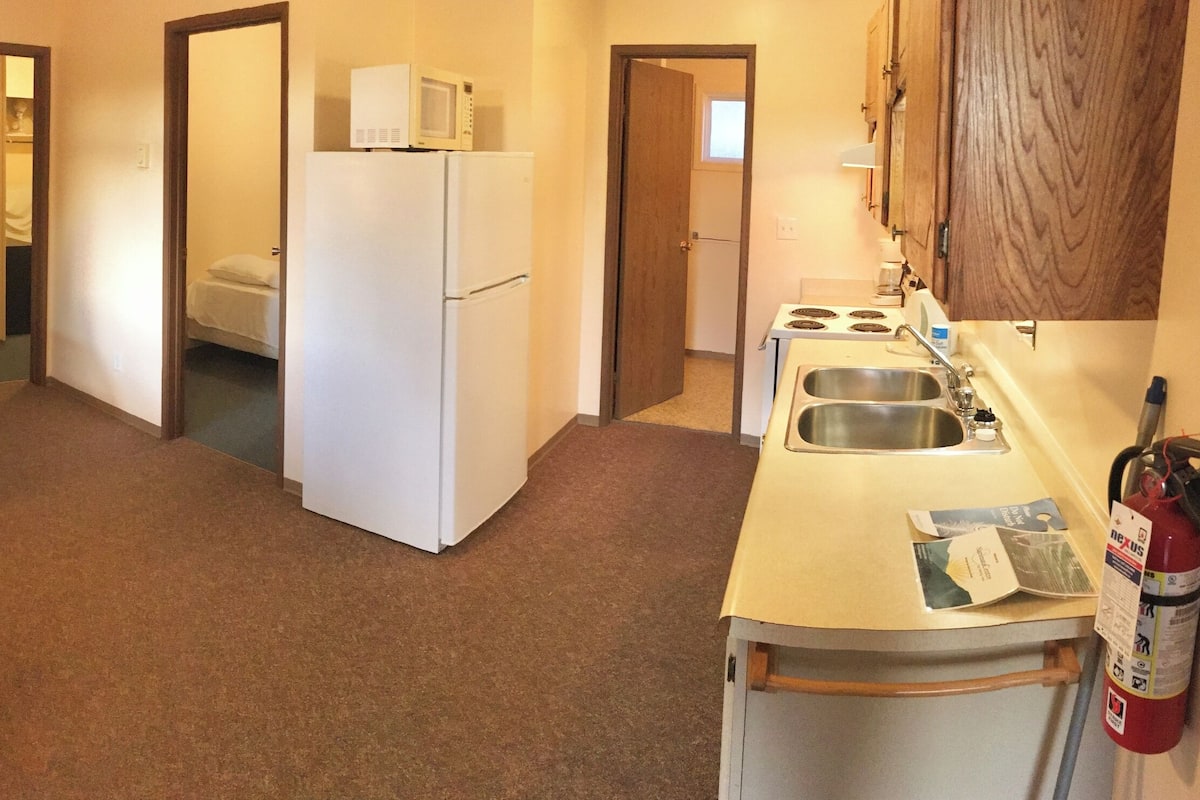 A compact kitchen with a refrigerator, microwave, stove, sink, and a fire extinguisher. There's a dining area with a table and chairs, and a small living space with a sofa and a window.