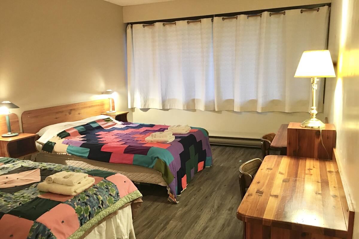 A hotel room with two beds, a wooden nightstand, a wooden chair, a desk, a lamp, and a window with curtains.