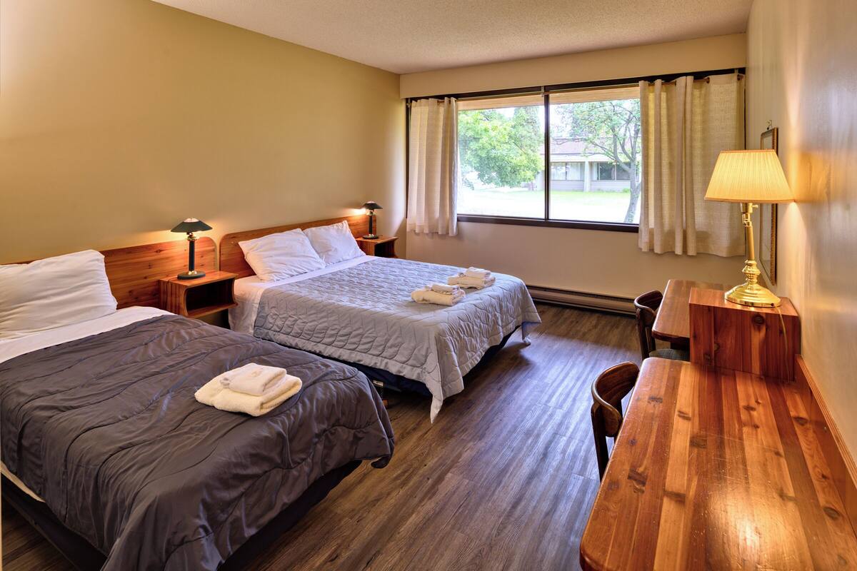 A hotel room with two beds, a wooden desk, a chair, a window with curtains, and a lamp.