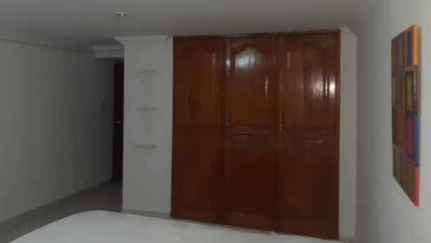 A bedroom with a wooden wardrobe, a white bed, and a colorful painting on the wall.