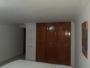 A bedroom with a wooden wardrobe, a white bed, and a colorful painting on the wall. - Habitacion Love, Hermosa Ubicada Cerca de Restaurantes Tipicos (Barranquilla)