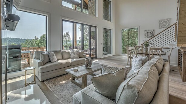 A spacious living room with a large sofa set, a coffee table, and a fireplace. There are large windows providing a view of the outdoors. - Juniper Ridge - Mountain Luxury | River & Golf Views | Outdoor Living and Hot Tub (Blue Ridge)