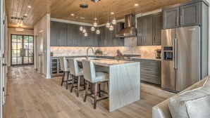A modern kitchen with dark grey cabinets, a stainless steel refrigerator, and a marble island with bar stools. - Juniper Ridge - Mountain Luxury | River & Golf Views | Outdoor Living and Hot Tub (Blue Ridge)