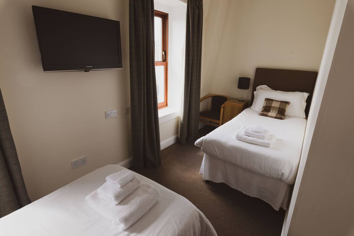 Ein Hotelzimmer mit zwei Betten, einem an der Wand befestigten Fernseher, einem Fenster mit Vorhängen und einem Nachttisch mit Lampe.