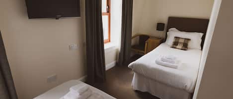 Een hotelkamer met twee bedden, een aan de muur bevestigde televisie, een raam met gordijnen, en een nachtkastje met een lamp.