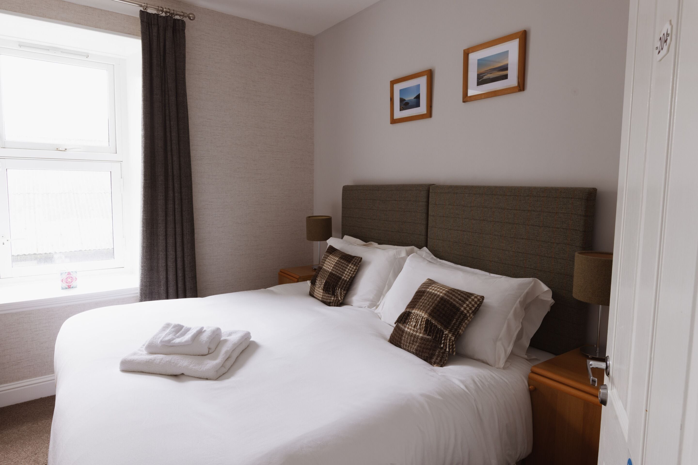 Una cama bien hecha con sábanas blancas y almohadas marrones, una lámpara de noche y una ventana con cortinas.