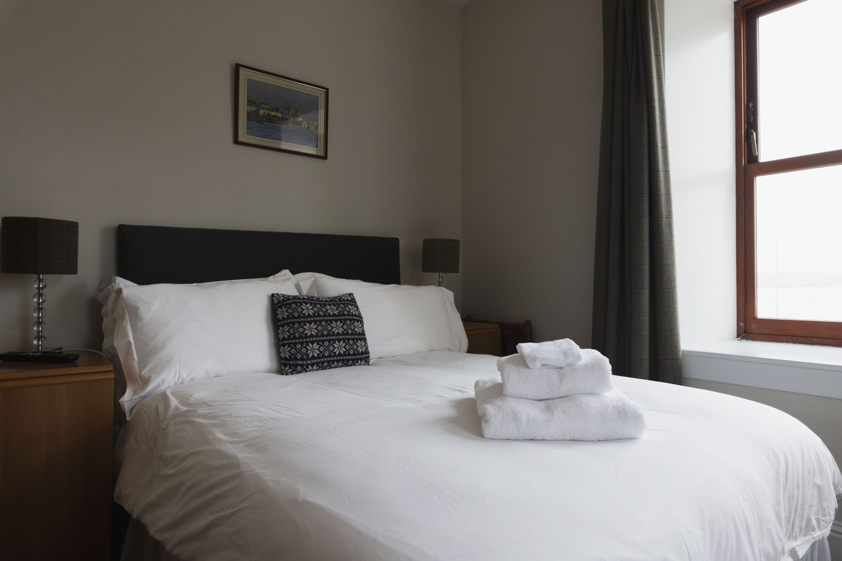 A neatly made bed with white linens and a folded towel, a dark headboard, two bedside lamps, a framed picture on the wall, and a window with curtains.