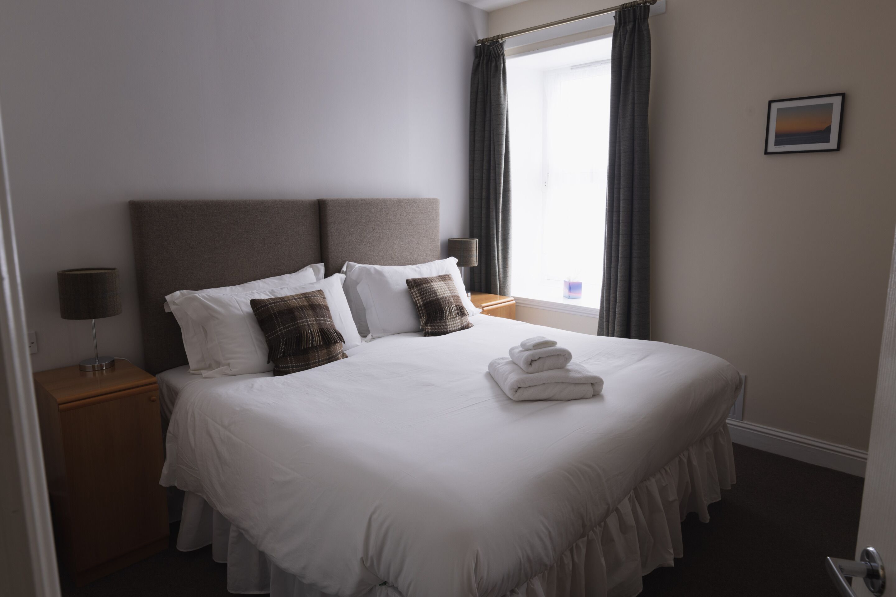Een net opgemaakt bed met witte lakens, een grijs hoofdbord en een bedlampje op een houten nachtkastje.