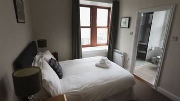 A hotel room with a large bed, a bedside lamp, a window with curtains, a radiator, and a bathroom visible through an open door.