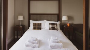 Una cama bien hecha con sábanas blancas, dos almohadas y dos toallas dobladas. Hay dos mesitas de noche, una con lámparas y una caja de pañuelos.