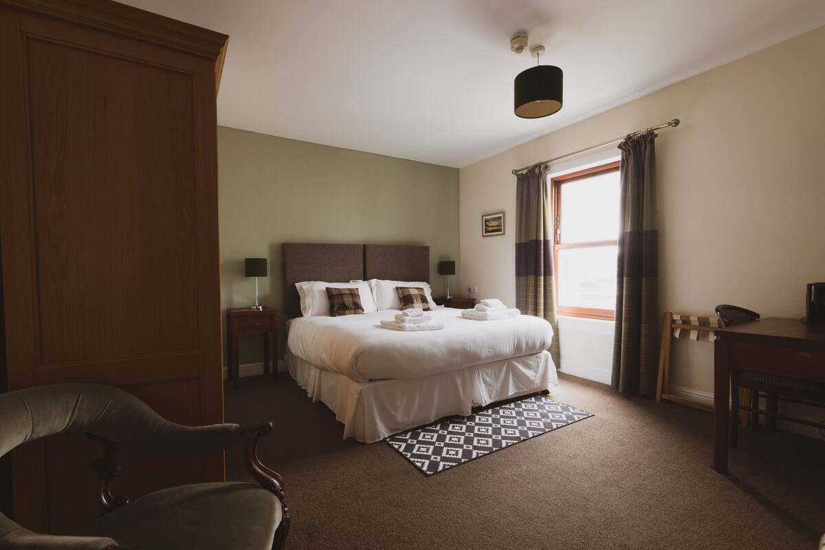 Ein Hotelzimmer mit einem großen Bett, einem Schreibtisch, einem Sessel und einem Fenster mit Vorhängen.
