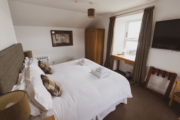A neatly made bed with white linens and a few decorative pillows, a wooden wardrobe, a television mounted on the wall, and a window with curtains. - Scalloway Hotel (Scalloway)