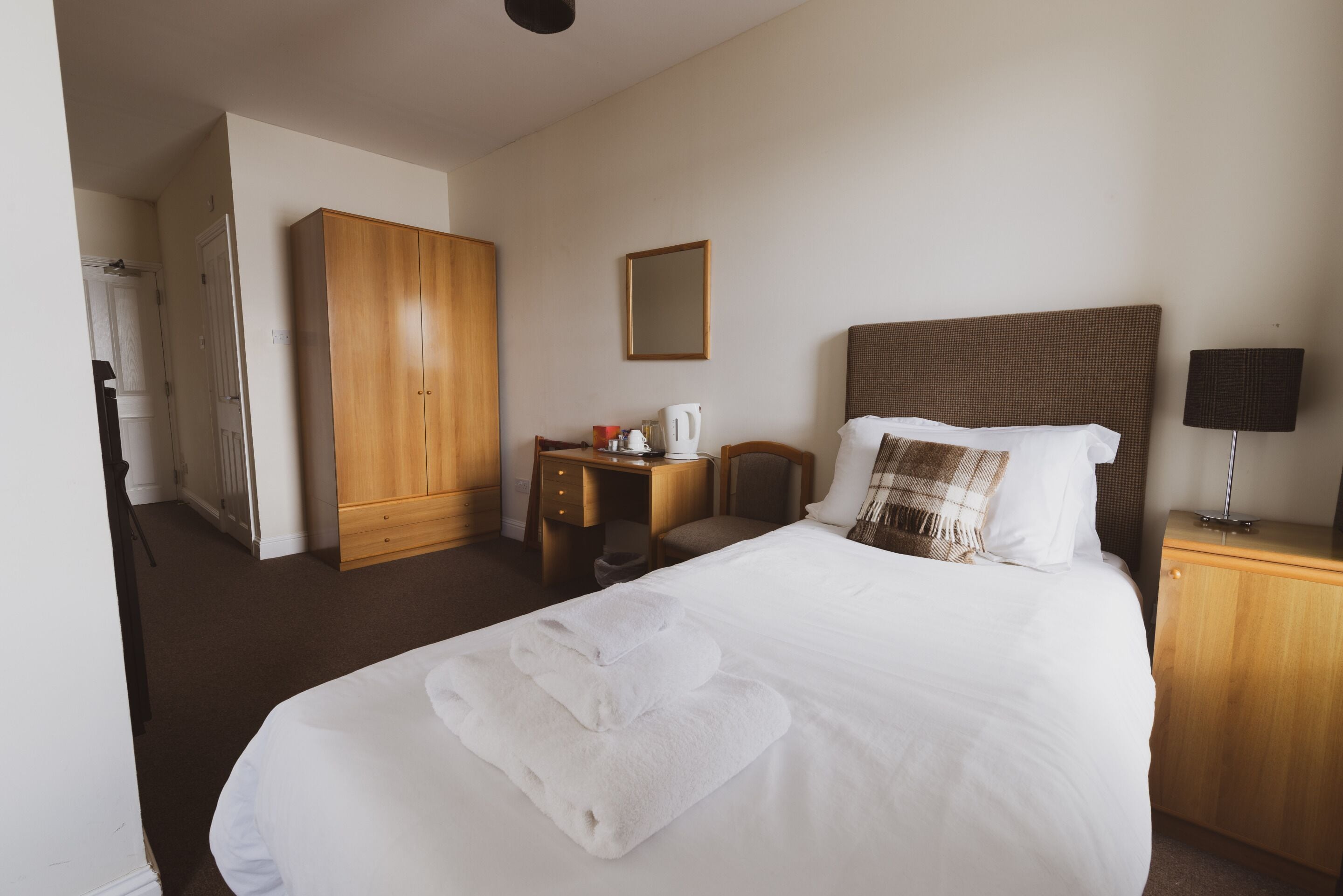 Una habitación de hotel con una cama grande, un armario de madera, un escritorio con silla, una mesita de noche con lámpara y un espejo en la pared.