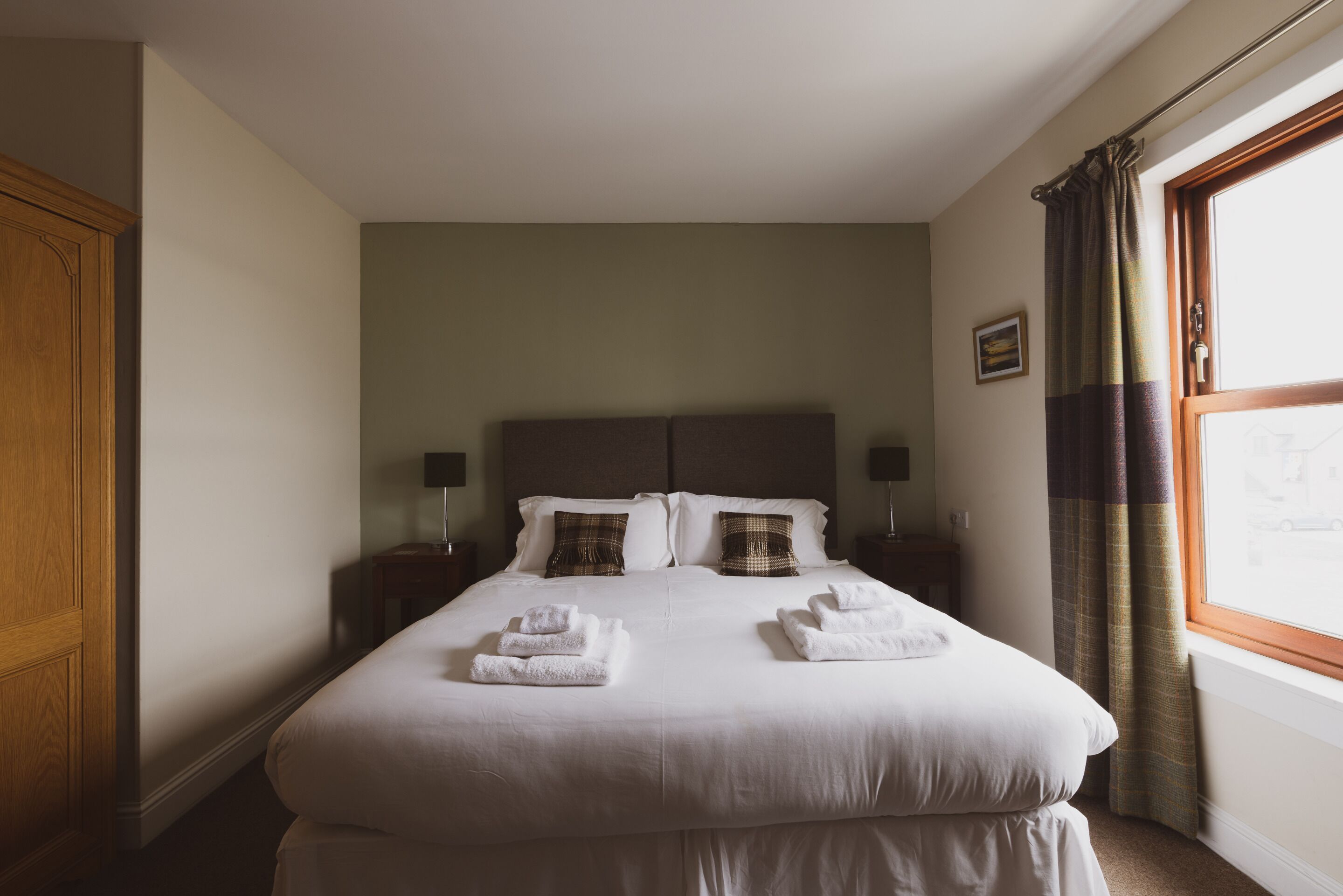 Una cama bien hecha con ropa de cama blanca y toallas dobladas, un armario de madera y una ventana con cortinas.