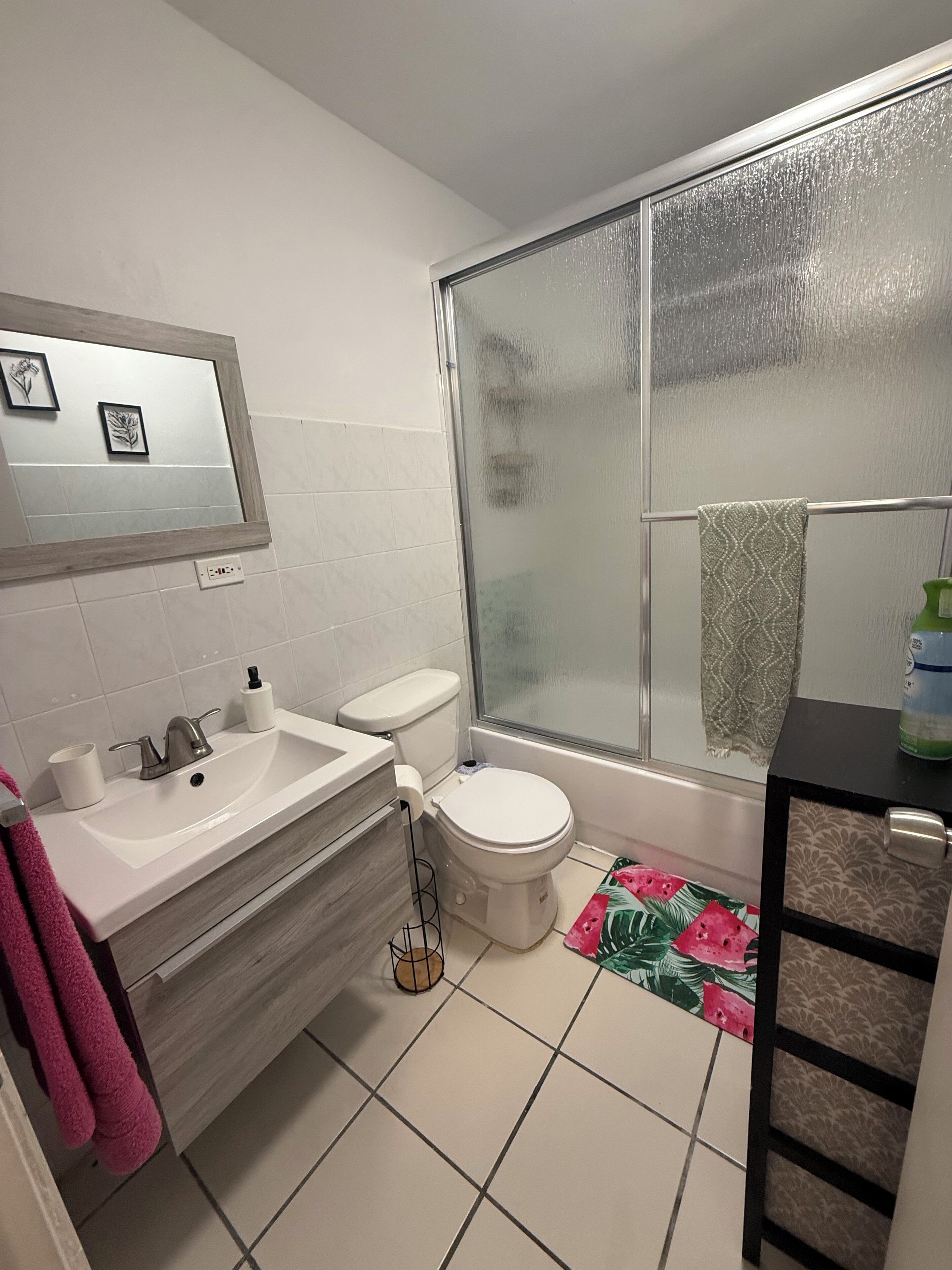 Et badeværelse med et hvidt håndvask, toilet og brusebad. Et spejl er monteret over håndvasken, og en håndklædestang har et håndklæde hængende på sig.