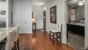 Une cuisine moderne avec des armoires blanches, un coin repas doté d’un comptoir en marbre et de chaises, et un couloir menant à une chambre à coucher équipée d’un lit et d’un ventilateur de plafond.
