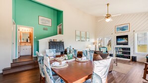 A dining area with a wooden table and chairs, a kitchen in the background, and a ceiling fan. - Walk to Beach! Boardwalk to Beach, Shared Pool, Pets OK! (Port Aransas)