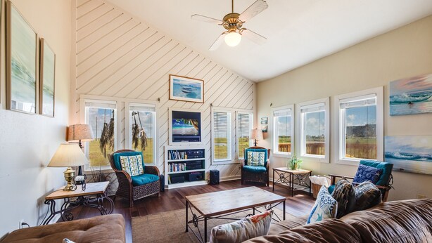 A spacious living room with a high ceiling, large windows, and a ceiling fan. There are multiple seating areas, including a sofa and chairs, with a bookshelf and a small table. - Walk to Beach! Boardwalk to Beach, Shared Pool, Pets OK! (Port Aransas)